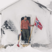 Mann ved hytta på snøfjell, med norsk flagg og Janus Wool Finse genser i farge rød