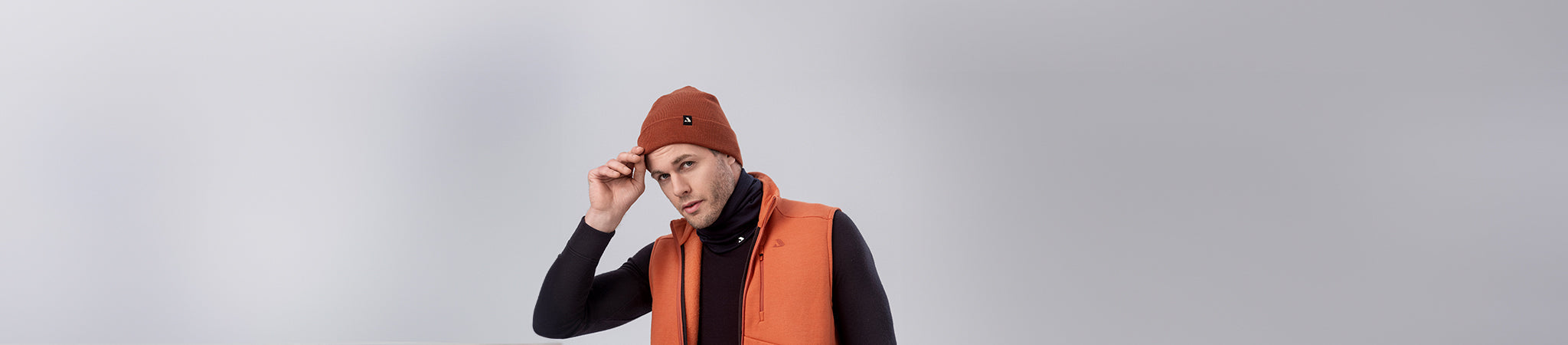 A man wearing an orange beanie and an orange vest over a black turtleneck stands against a plain light gray background, looking at the camera and adjusting his beanie.