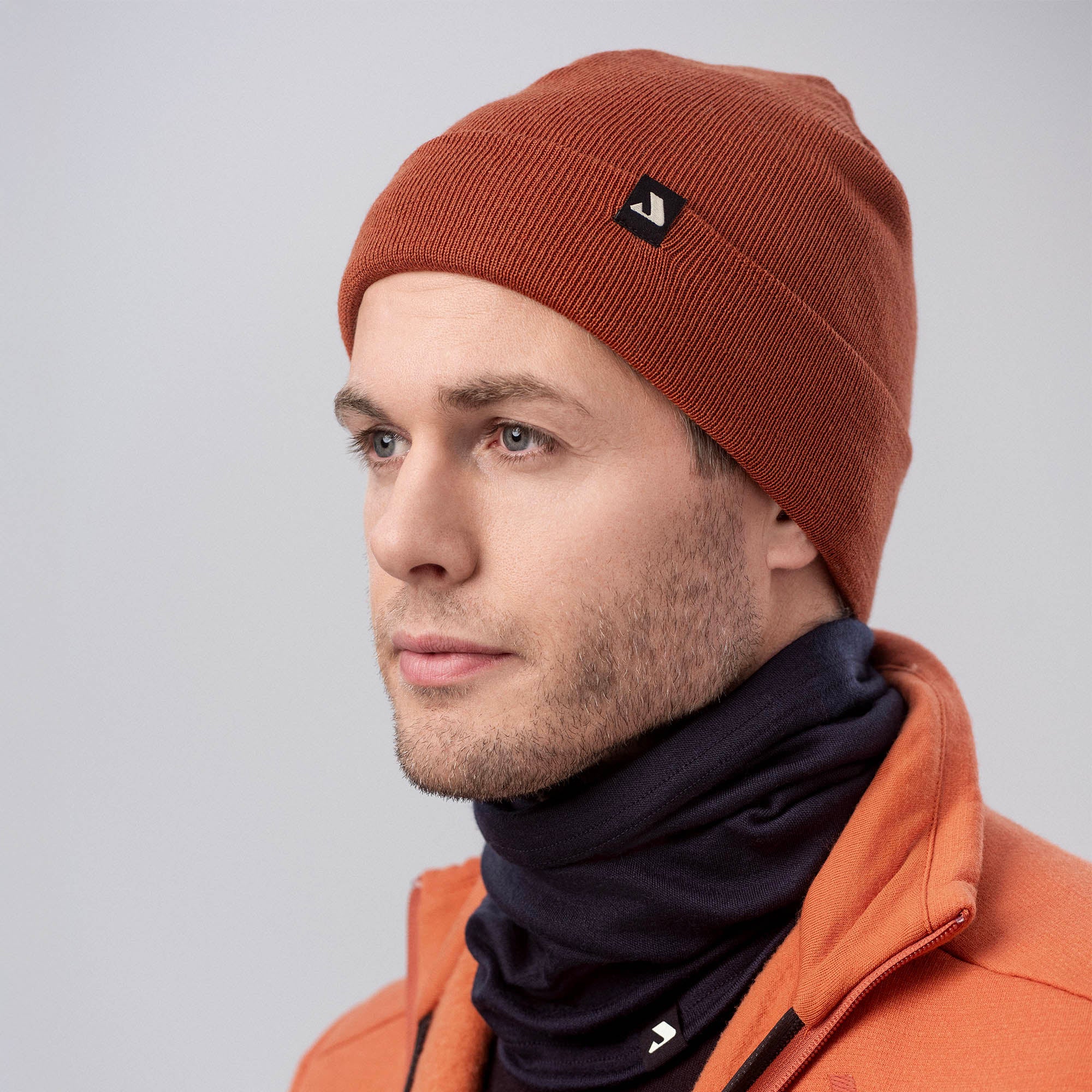 A man wearing a MeshWool beanie, an orange jacket, and a black neck gaiter looks off to the side against a light gray background.