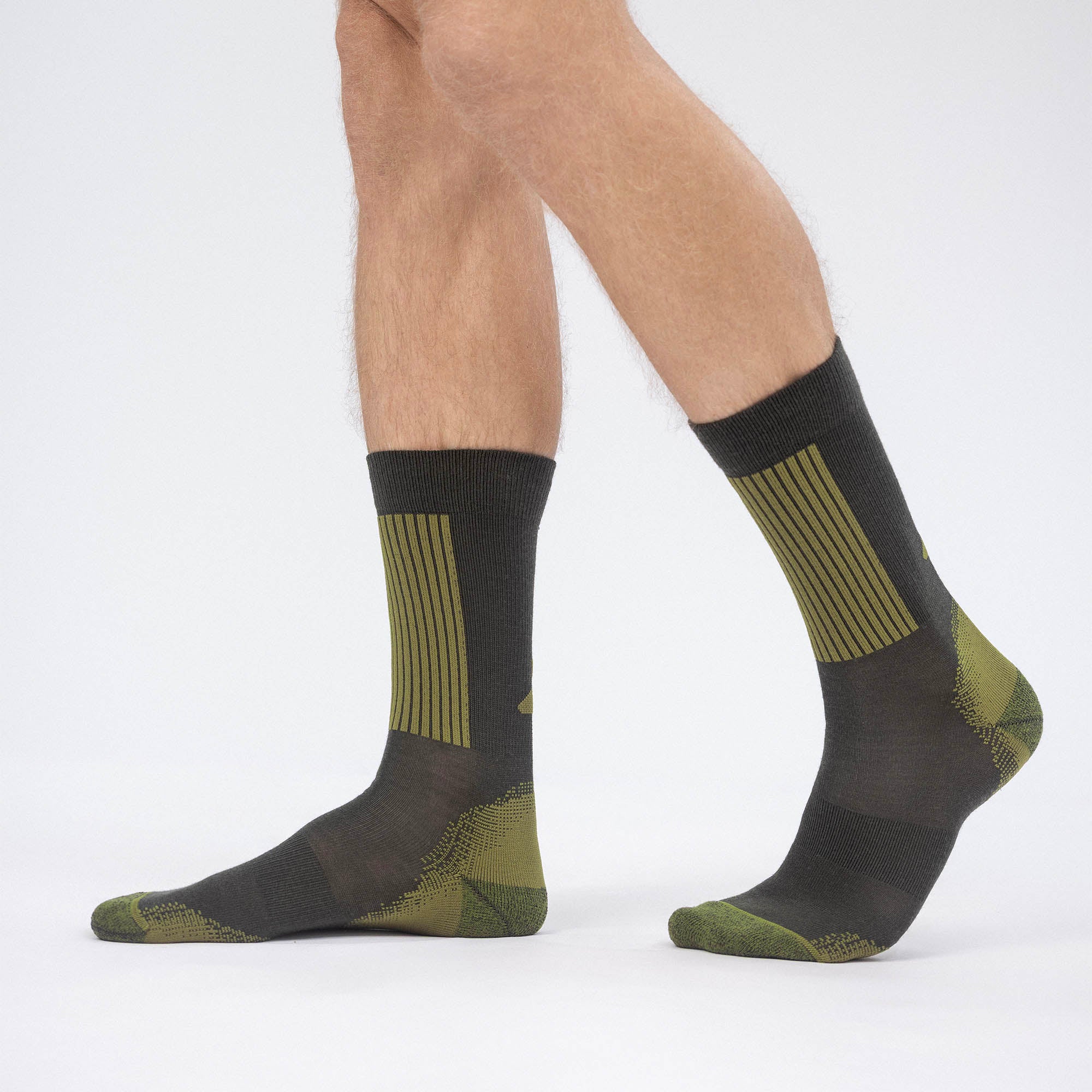A person wearing Socks Merino – Hiking in green and black with geometric patterns stands on a white background, showing only the lower legs and feet to highlight hiking comfort.