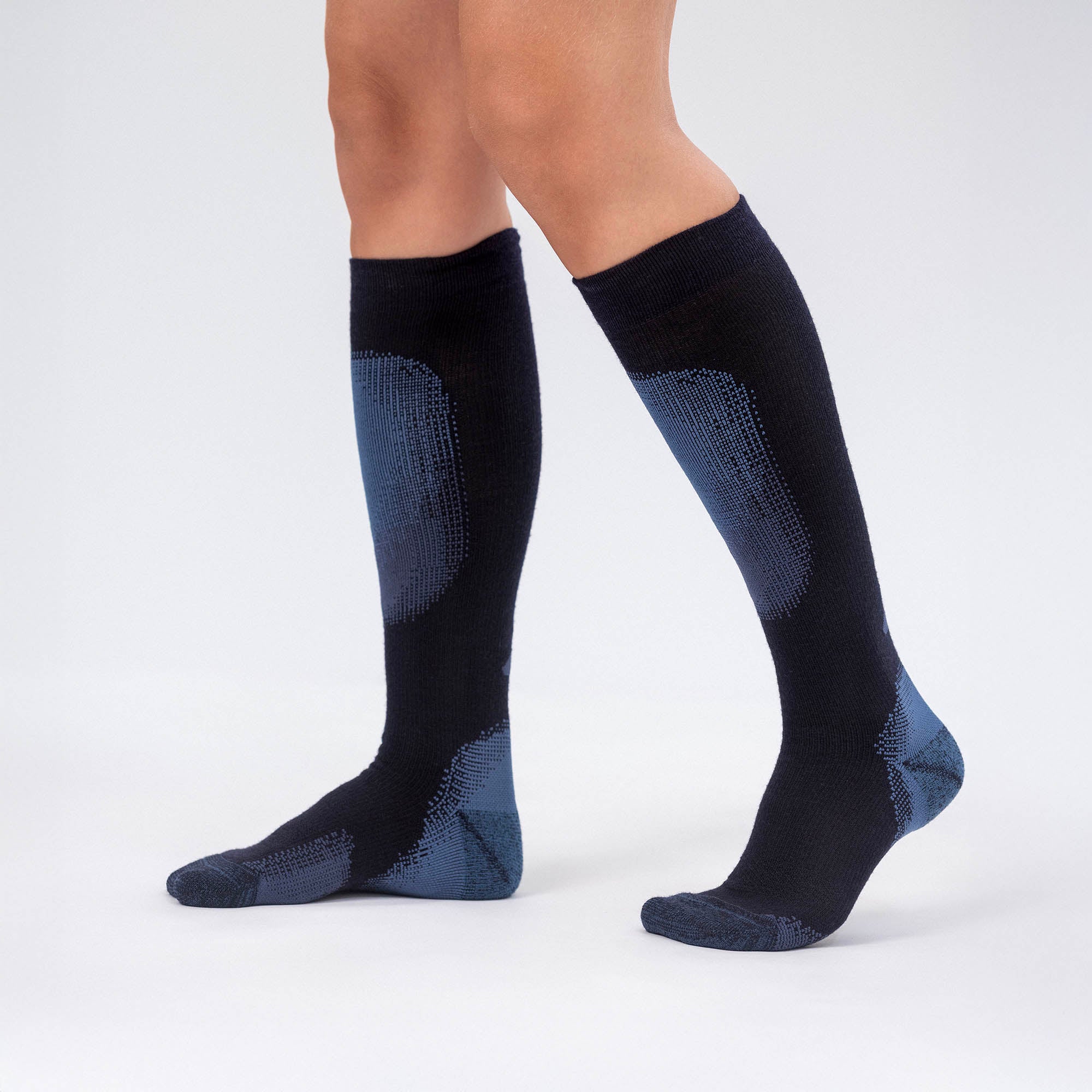 A person wearing Socks Merino – Alpine, knee-high black with blue accents, stands on a plain light background, showing only their legs and feet—ideal for slipping into ski boots after alpine adventures.