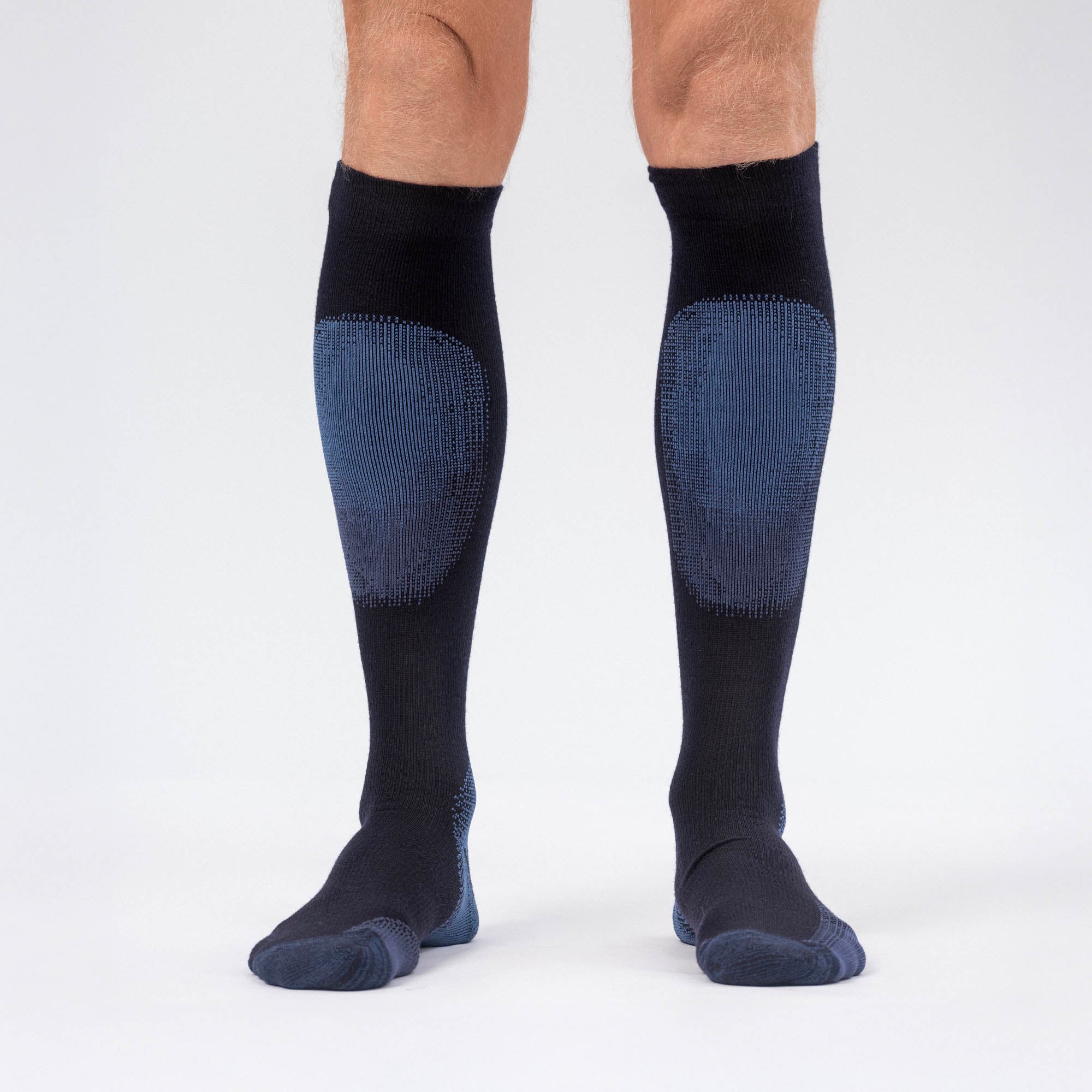 Wearing Socks Merino – Alpine, knee-high black Merino wool socks with blue padding on the shins and toes for alpine skiing, a person's lower legs and feet are visible against a white background.