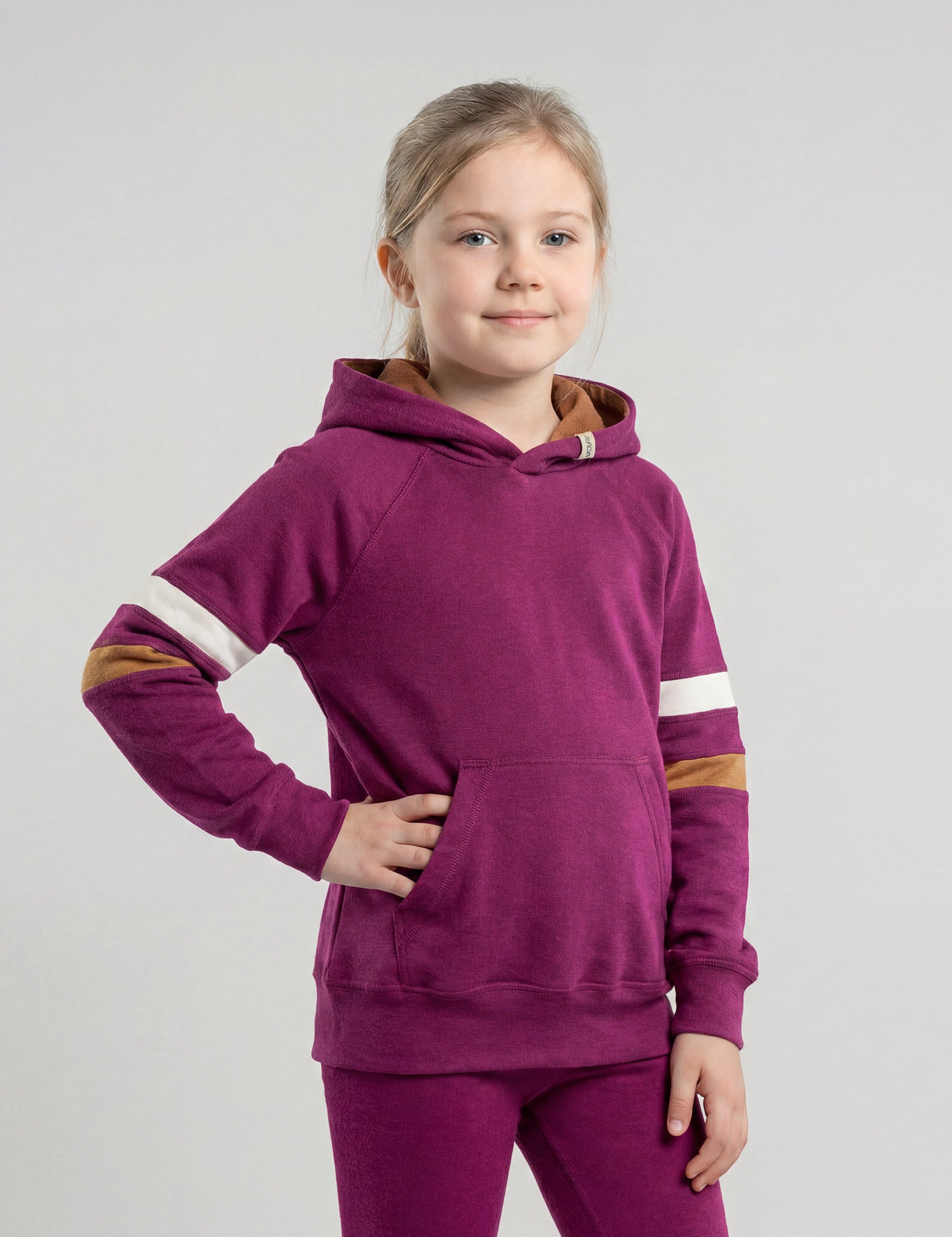 A young girl with blonde hair stands with one hand on her hip, smiling at the camera while wearing the Hoodie merino - Janus Go 250 Kids, featuring white and brown striped sleeves, against a plain light gray background.
