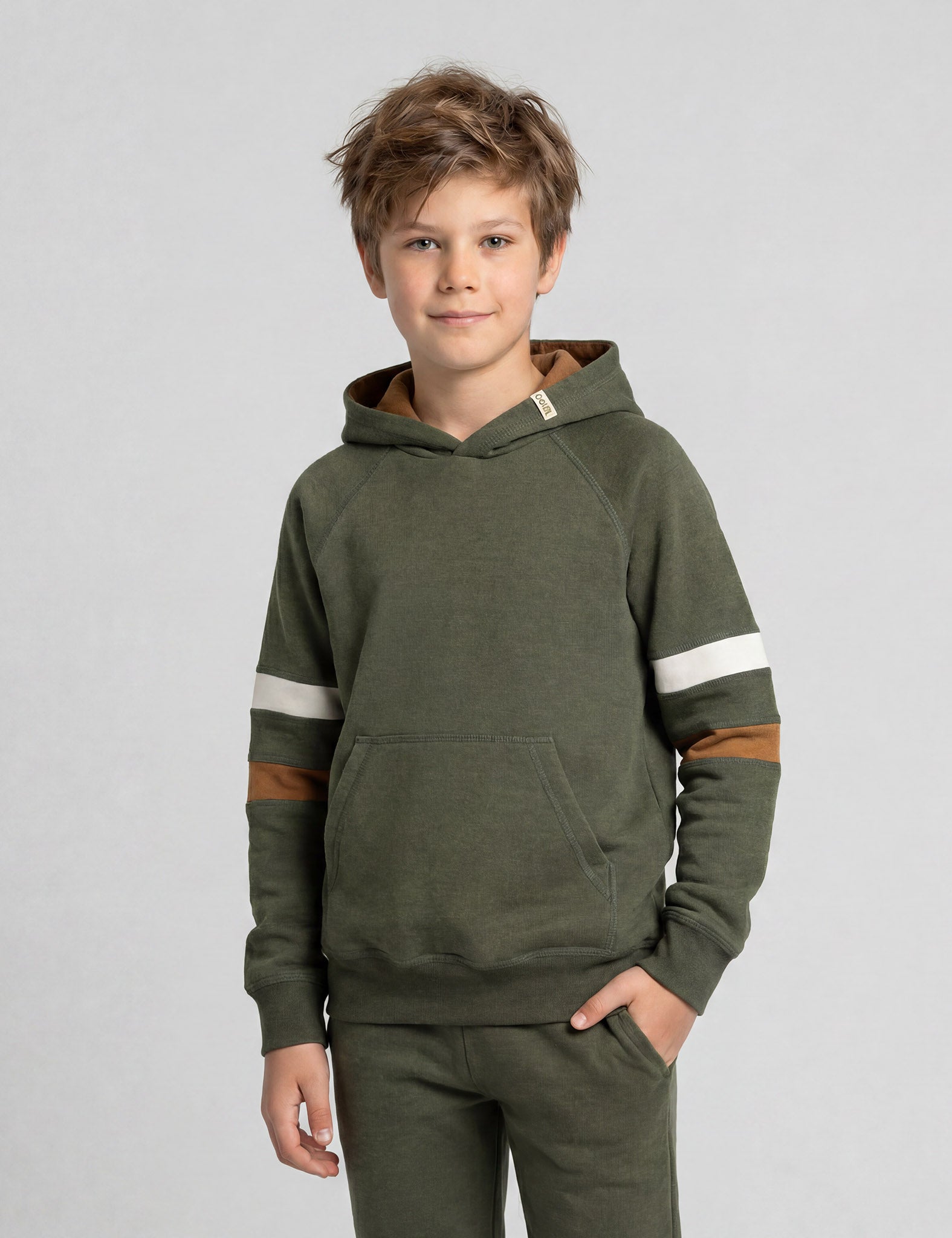 A young boy with tousled light brown hair wears the Janus Go 250 Kids Hoodie merino in olive green with white and brown sleeve stripes and matching pants, standing against a plain light gray background.