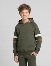 A young boy with tousled light brown hair wears the Janus Go 250 Kids Hoodie merino in olive green with white and brown sleeve stripes and matching pants, standing against a plain light gray background.