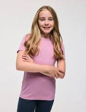 A smiling young girl with long blonde hair wears the T-Shirt – Merino Kids Light in Mauve Orchid and dark pants, standing against a plain white background with her arms casually crossed for all-day comfort.