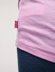 Close-up of a person wearing the T-Shirt – Merino Kids Light in light pink, featuring a small Norwegian flag tag on the side seam and paired with dark blue pants for all-day comfort.