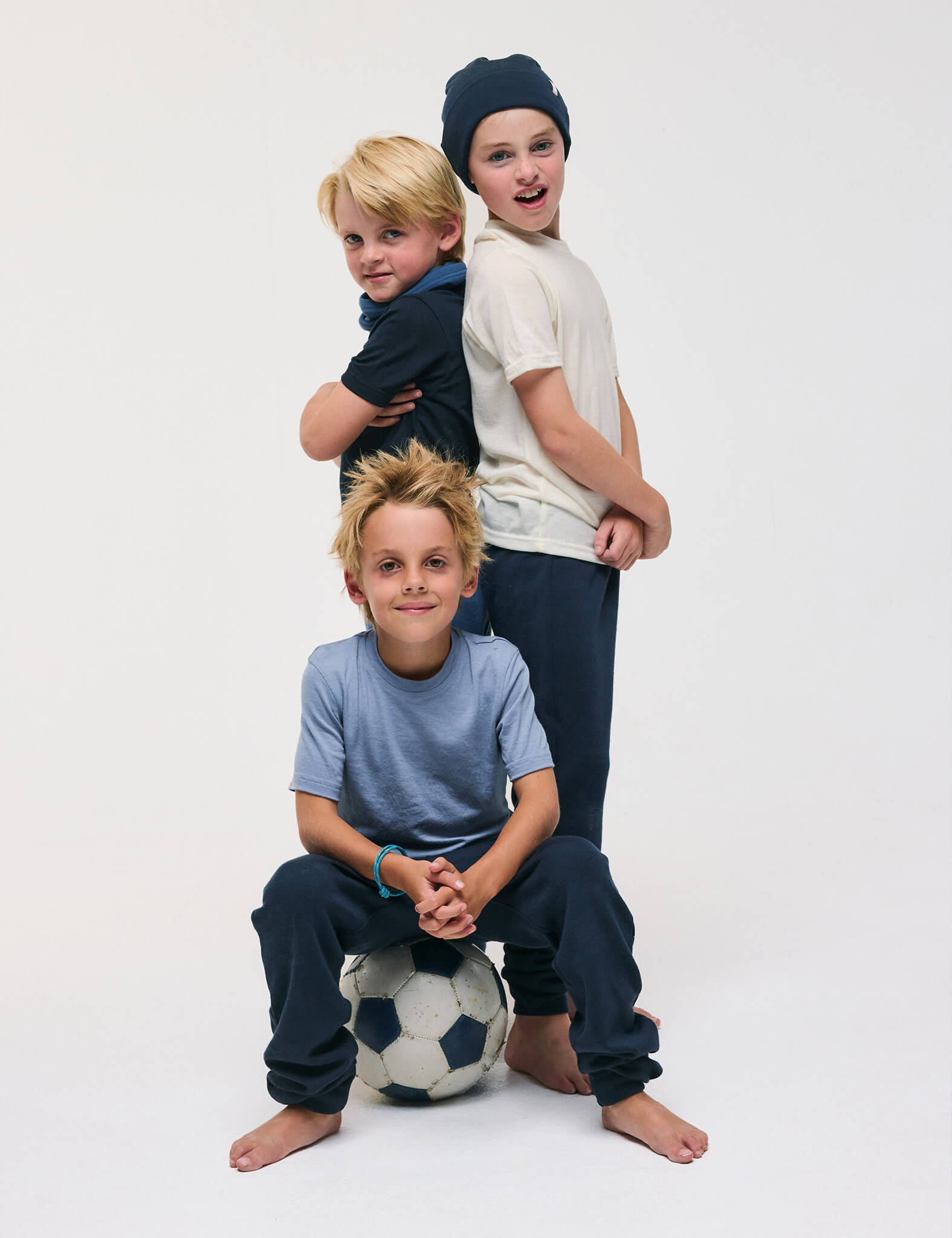 Three barefoot children pose in casual clothes. Two stand back-to-back while the third sits cross-legged on the floor with hands resting on a soccer ball, all smiling against a plain white background. Their T-Shirt – Merino Kids Light tops provide all-day comfort and superior temperature regulation as they play.