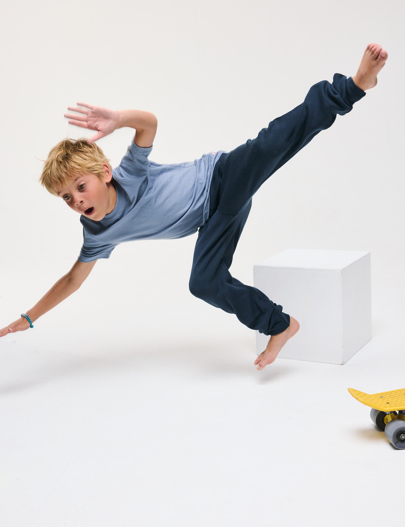 A young boy wearing the T-Shirt – Merino Kids Light and blue pants is falling through the air with arms outstretched, near a white cube and a yellow skateboard on the floor.