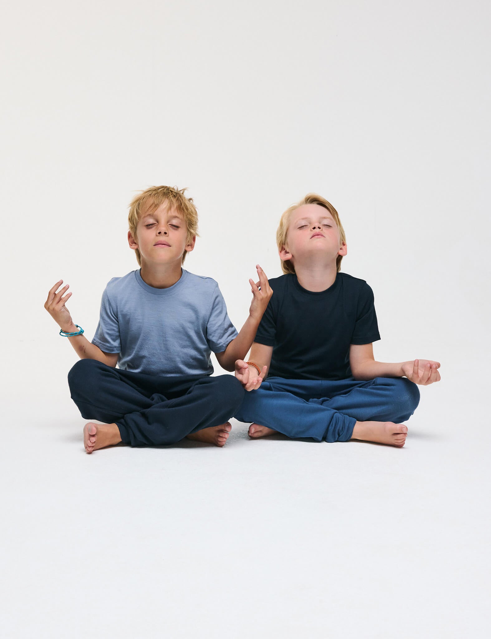 Two young boys sit cross-legged on the floor, eyes closed and hands in meditation poses, appearing calm and relaxed against a plain white background. Their T-Shirt – Merino Kids Light provides all-day comfort and natural temperature regulation, enhancing their peaceful experience.