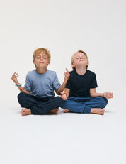 Two young boys sit cross-legged on the floor, eyes closed and hands in meditation poses, appearing calm and relaxed against a plain white background. Their T-Shirt – Merino Kids Light provides all-day comfort and natural temperature regulation, enhancing their peaceful experience.