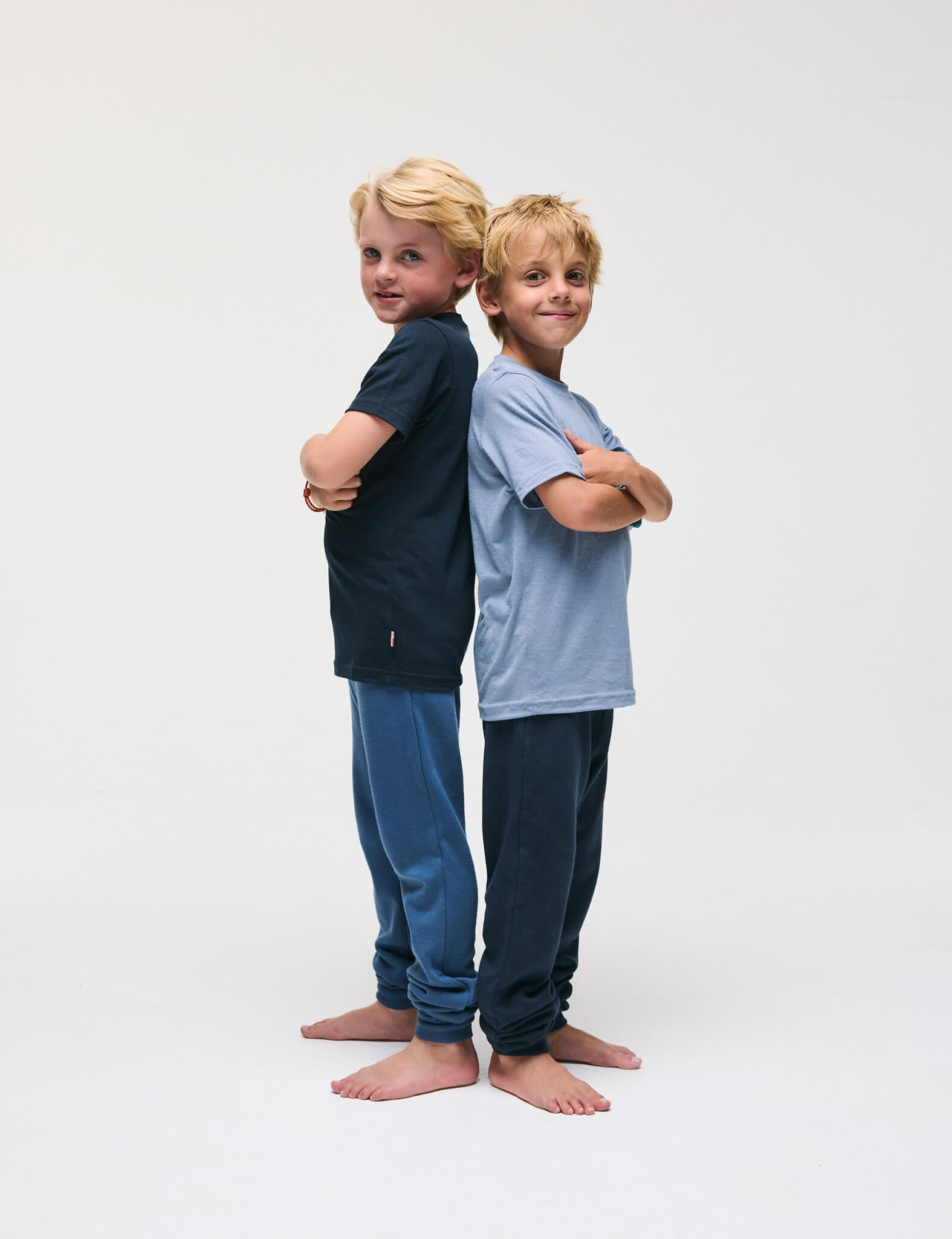 Two young boys stand back to back with arms crossed, both barefoot and smiling. One wears a dark shirt and blue pants; the other wears a light blue T-Shirt – Merino Kids Light and dark pants. Their outfits are designed for all-day comfort, featuring materials that provide excellent temperature regulation. The background is plain white.