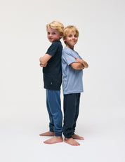 Two young boys stand back to back with arms crossed, both barefoot and smiling. One wears a dark shirt and blue pants; the other wears a light blue T-Shirt – Merino Kids Light and dark pants. Their outfits are designed for all-day comfort, featuring materials that provide excellent temperature regulation. The background is plain white.