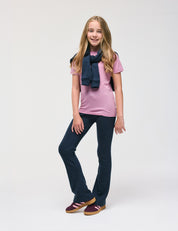 A young girl with long blonde hair is standing and smiling against a plain white background, wearing the Merino Kids Light T-Shirt in pink, paired with a navy sweater draped over her shoulders, navy pants, and maroon shoes. This outfit offers all-day comfort and excellent temperature regulation.