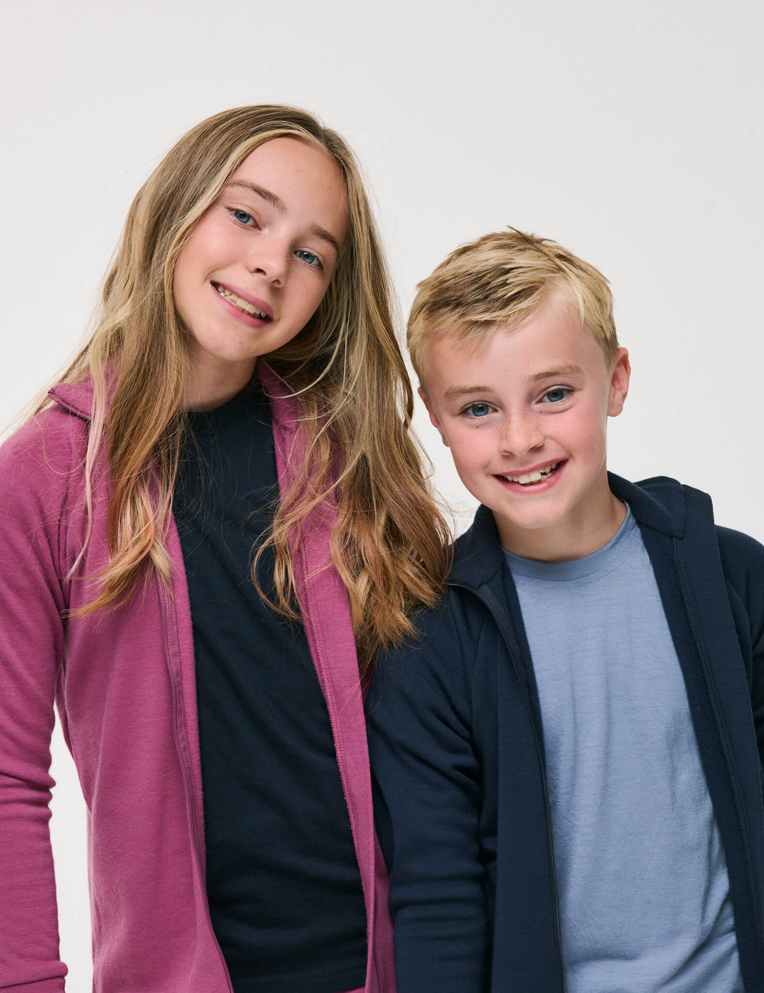 Two smiling children, a girl in a pink hoodie and a boy in a navy hoodie layered over the T-Shirt – Merino Kids Light in light blue, stand close together against a plain white background, showcasing all-day comfort and excellent temperature regulation.