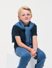 A young boy with blond hair sits on a white block, hands clasped in his lap and a slight smile, wearing the T-Shirt – Merino Kids Light in navy for all-day comfort, along with blue pants and a blue sweatshirt draped over his shoulders.