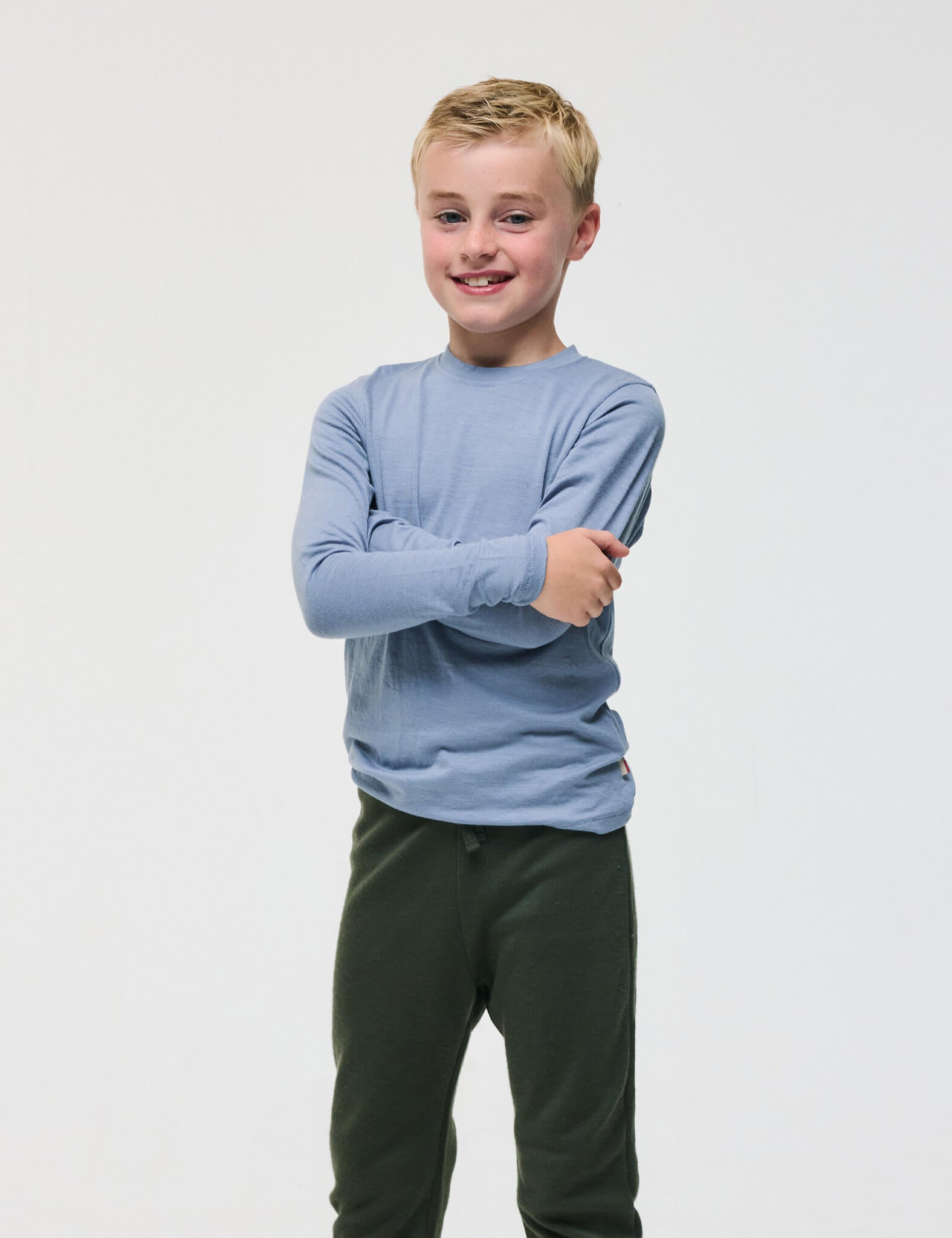 A smiling young boy with short blonde hair stands with arms crossed, wearing the Crewneck – Merino Kids Light long-sleeve shirt in light blue and dark green pants against a plain white background.