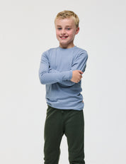 A smiling young boy with short blonde hair stands with arms crossed, wearing the Crewneck – Merino Kids Light long-sleeve shirt in light blue and dark green pants against a plain white background.
