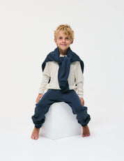 A young boy with messy blonde hair sits barefoot on a white cube, wearing the Crewneck – Merino Kids Light long-sleeve shirt in soft cream and navy sweatpants, with a navy sweatshirt tied around his shoulders, against a plain white background.