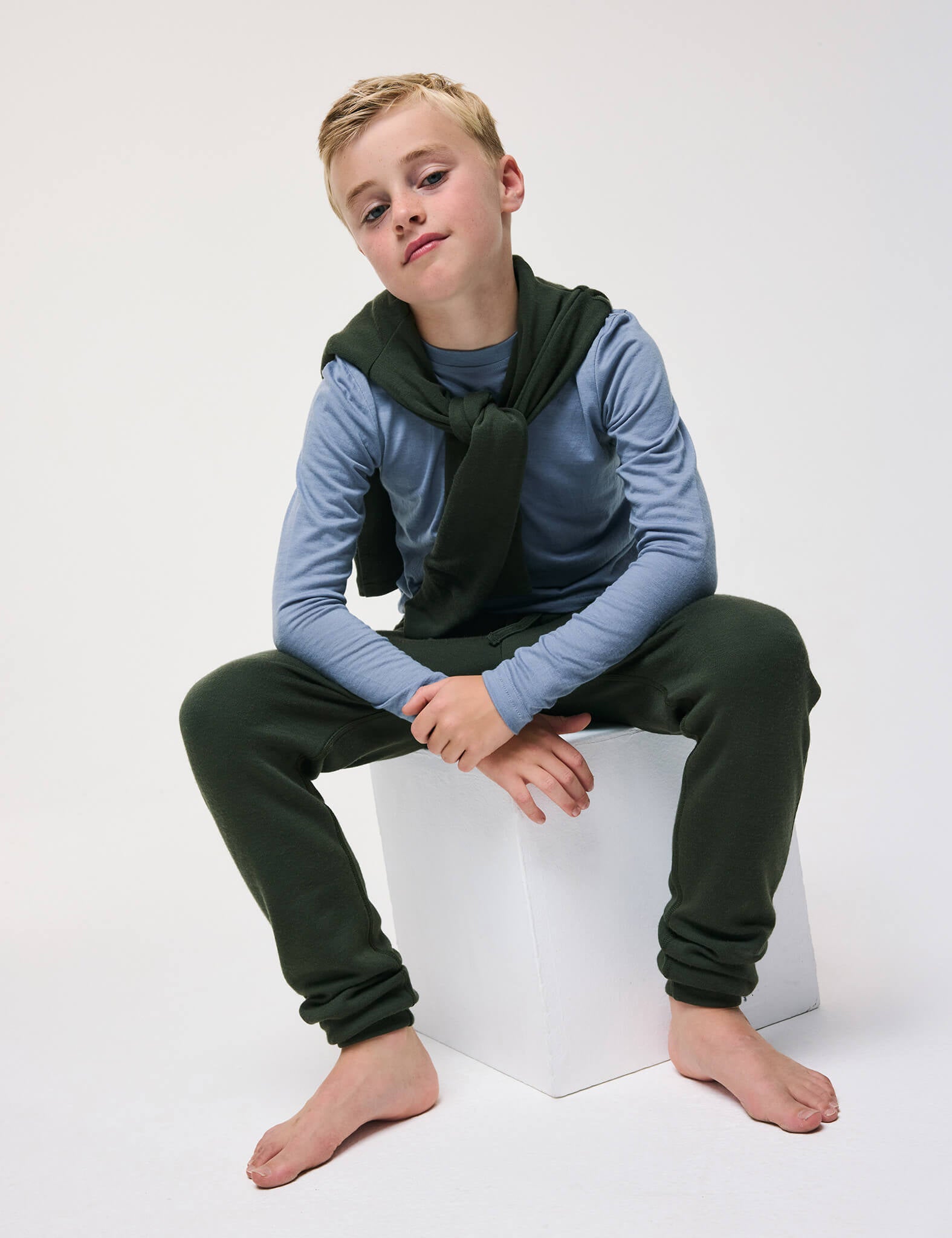 A young boy with blonde hair sits barefoot on a white cube, confidently modeling the Crewneck – Merino Kids Light in Troposphere, styled with dark green pants and a soft green sweater draped over his shoulders against a plain backdrop.