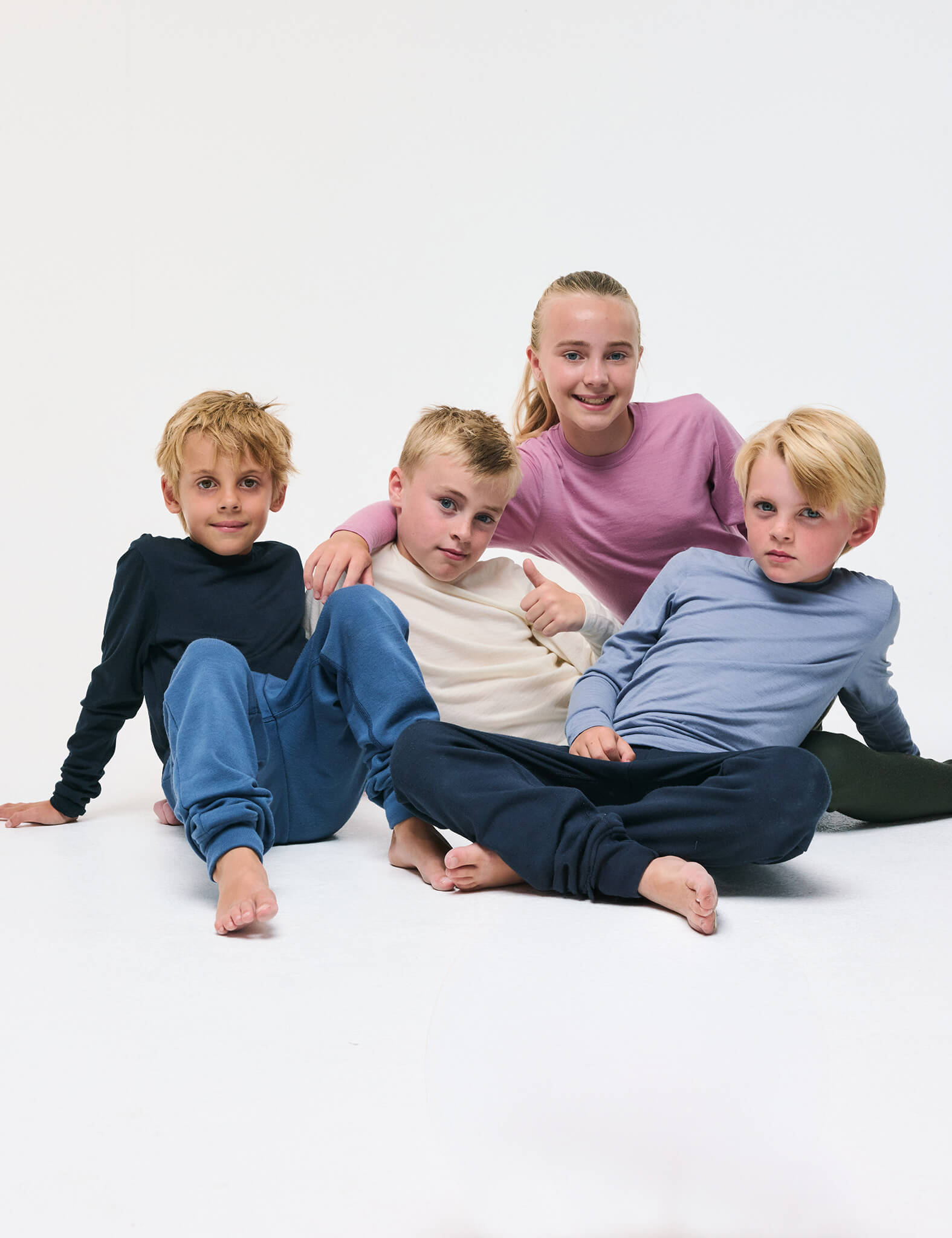 Four children sit together on the floor, smiling and wearing cozy Crewneck – Merino Kids Light long-sleeve shirts and sweatpants in blue, cream, pink, and navy tones, against a plain white background.