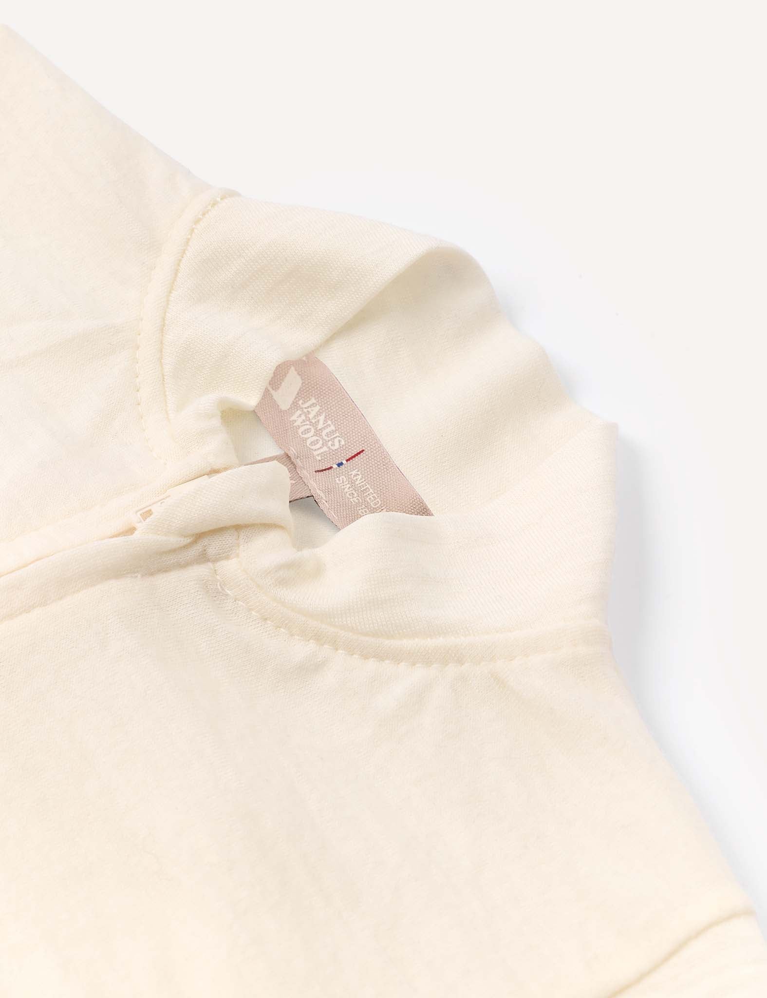A close-up of the Pyjamas – Merino Baby Light showcases its cream-colored fabric, featuring a collar, partial zipper, and a beige inner tag with text. The material appears soft and breathable, hinting at the comfort these pyjamas provide. The background is white.
