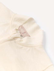 A close-up of the Pyjamas – Merino Baby Light showcases its cream-colored fabric, featuring a collar, partial zipper, and a beige inner tag with text. The material appears soft and breathable, hinting at the comfort these pyjamas provide. The background is white.