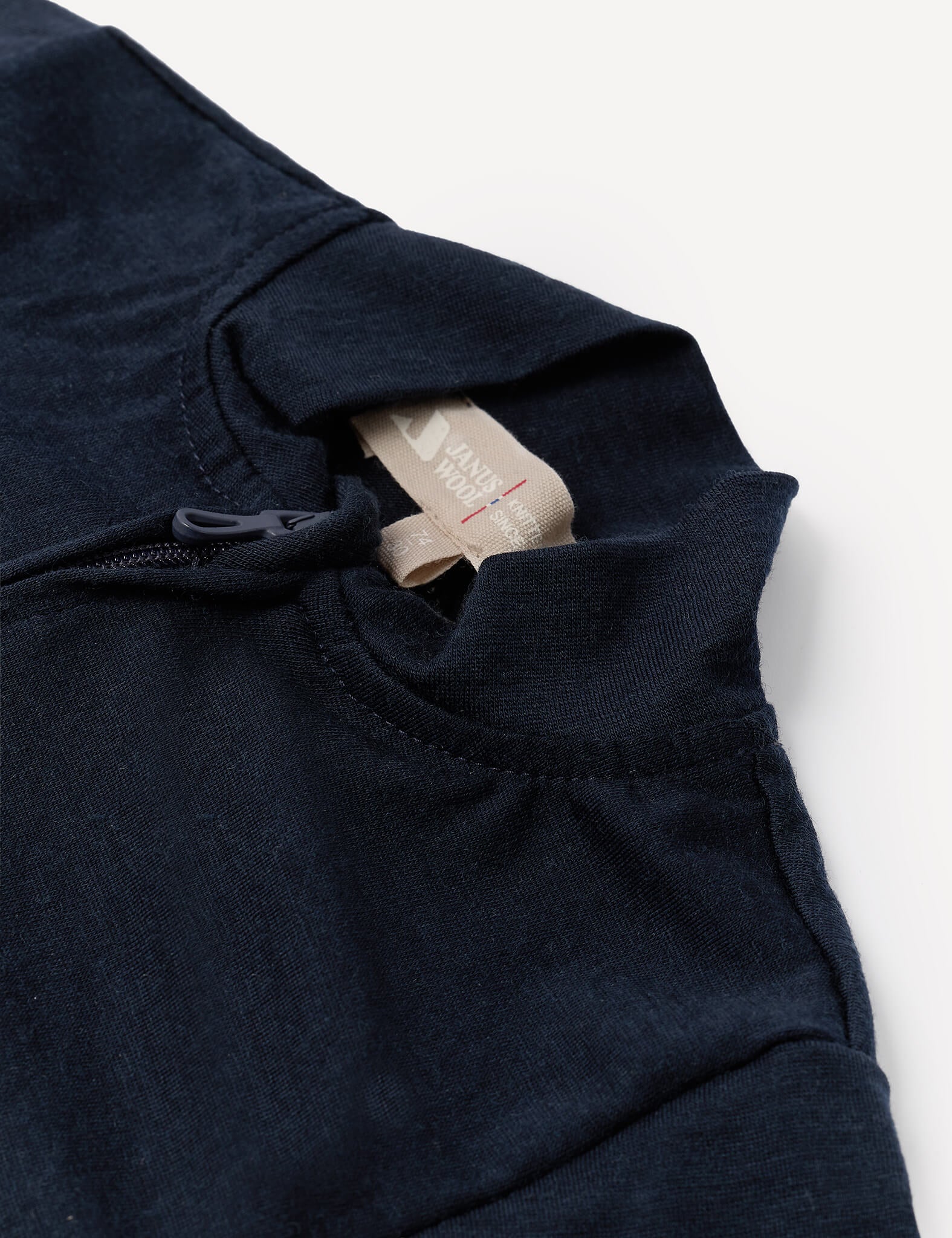 Close-up of the Pyjamas – Merino Baby Light in navy blue, featuring a standing collar with an exposed zipper and a beige tag with red text at the neckline, laid on a white surface. Crafted from breathable merino nightwear material designed for superior comfort.