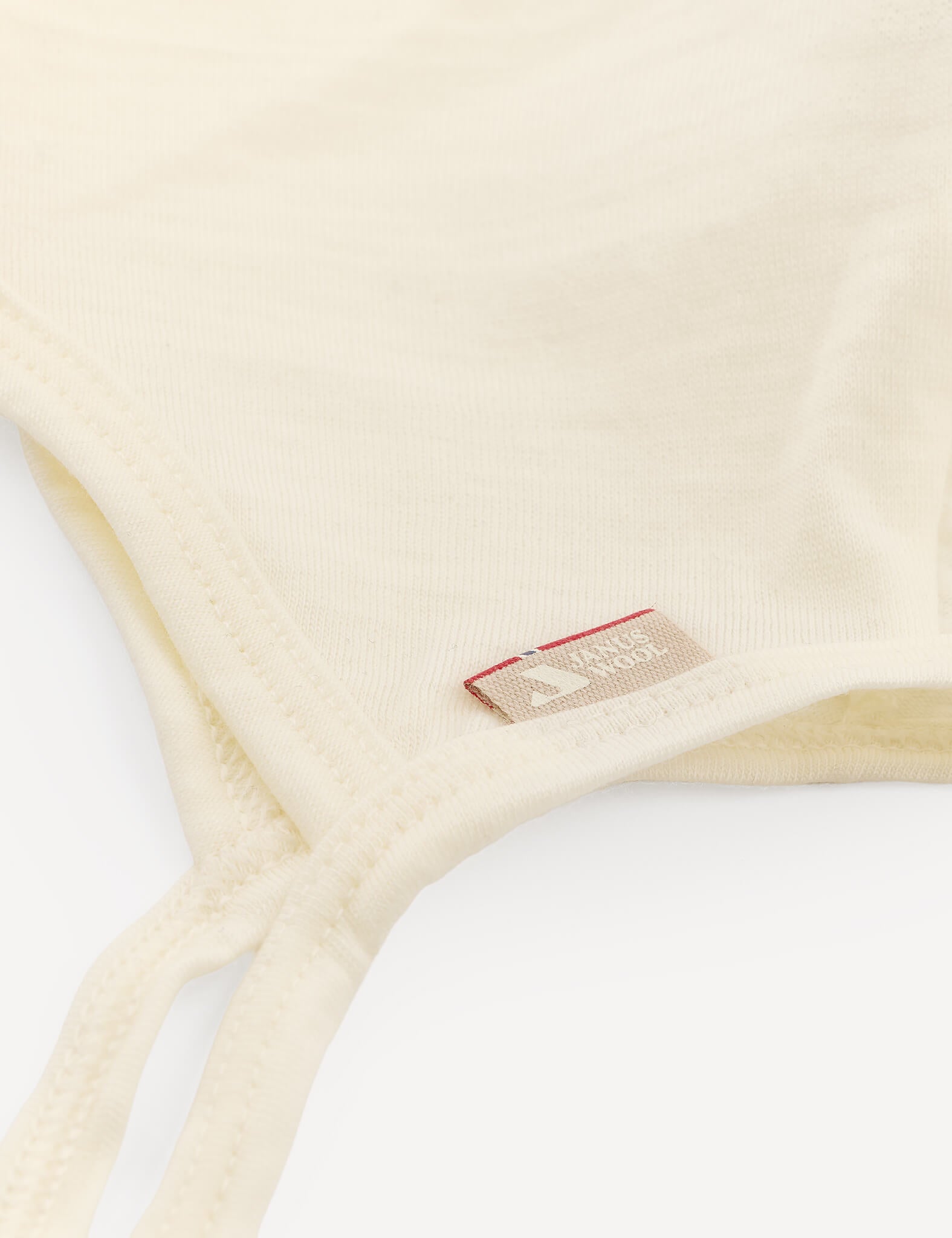 Close-up of the Bonnet – Merino Baby Light in a cream shade, showing a small fabric tag with text and a red accent. The soft, lightweight merino wool fabric is similar to materials used in breathable baby hats and bonnets.