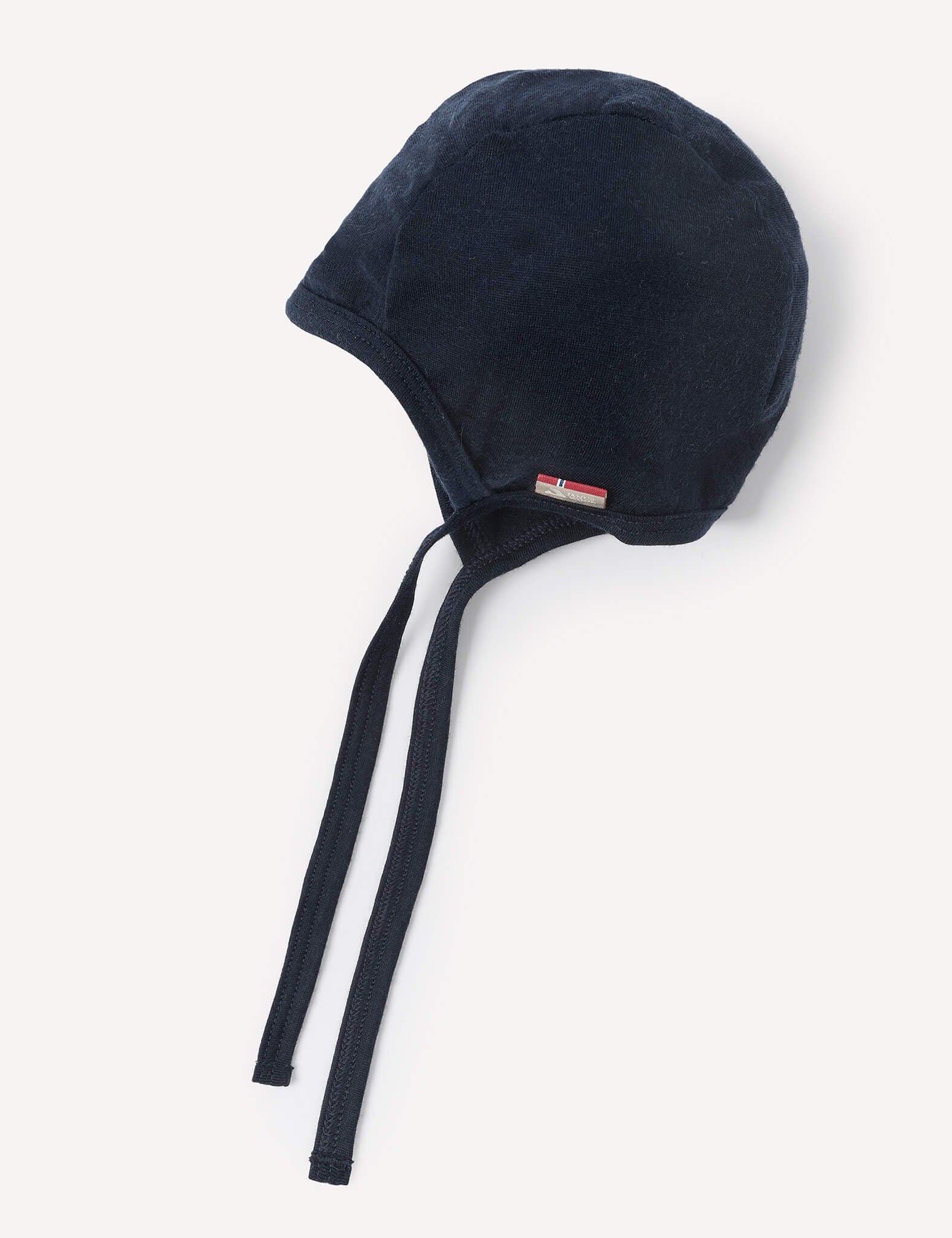 A navy Merino Baby Light bonnet with chin ties, displayed against a plain white background. A small rectangular tag with a red and white design is attached near the edge.