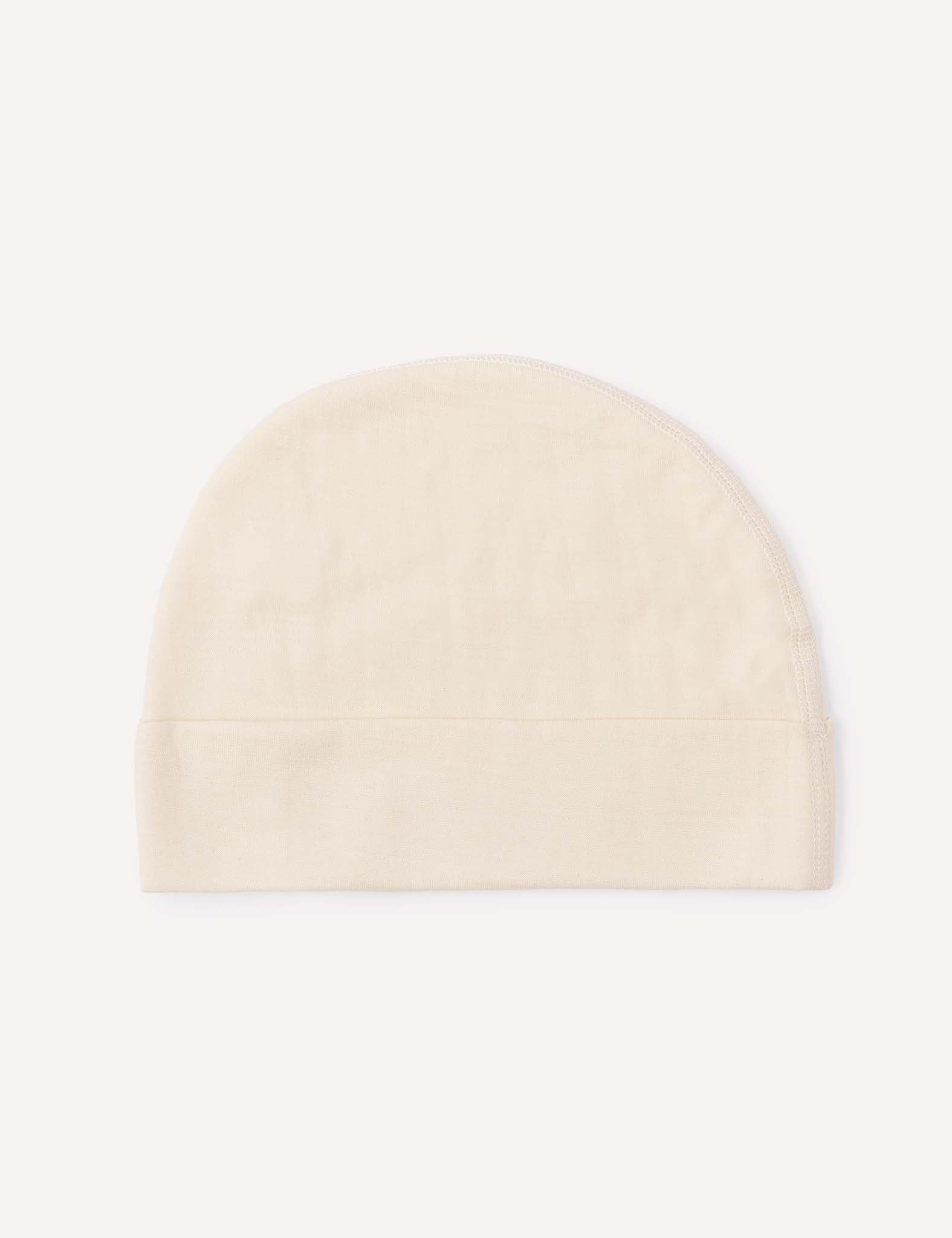 The Beanie – Merino Baby Light, in a simple light beige shade, features a rounded top and a fold-over brim. It is displayed flat against a white background, highlighting its breathable design.
