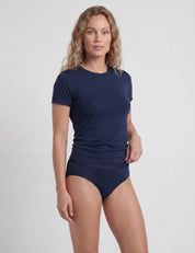 A woman with wavy light brown hair wears a navy blue Classic Merino 160 Women Briefs and matching t-shirt, standing against a plain light gray background. She is facing slightly to the side with a neutral expression.
