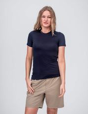 A woman with long, light brown hair wears the T-shirt Active – Classic Merino 160 Women in Navy and tan shorts, standing against a plain light gray background. She faces the camera neutrally, showcasing the shirt’s temperature-regulating qualities.