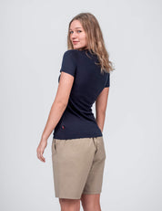 A woman with light brown hair wears the T-shirt Active – Classic Merino 160 Women in navy, paired with beige knee-length shorts. She stands turned away, smiling back over her shoulder against a plain light gray background.