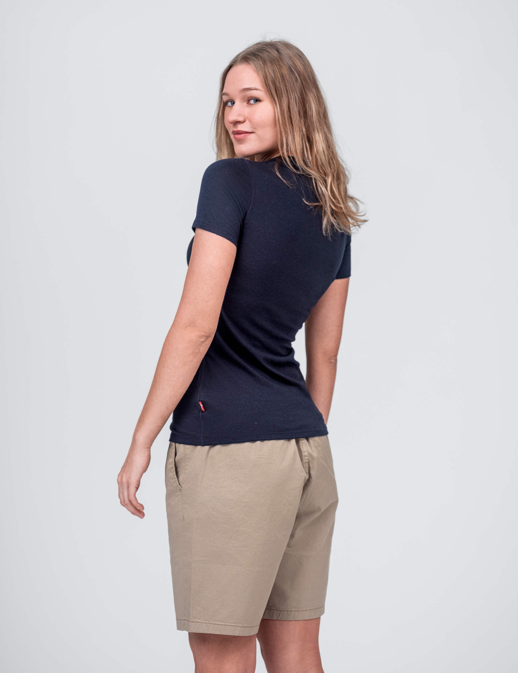A woman with light brown hair wears the T-shirt Active – Classic Merino 160 Women in navy, paired with beige knee-length shorts. She stands turned away, smiling back over her shoulder against a plain light gray background.