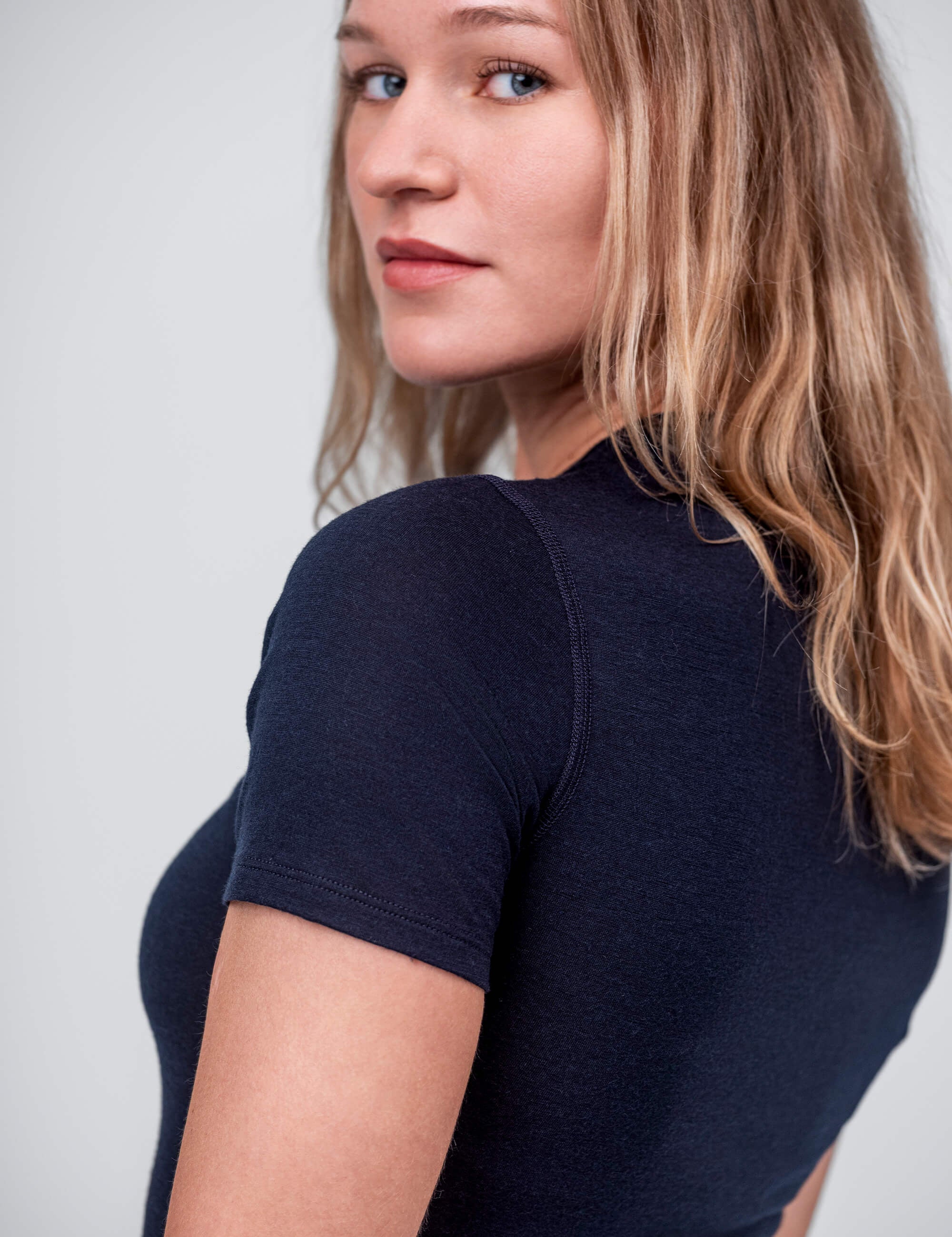 A woman with long, blonde hair and blue eyes wears the T-shirt Active – Classic Merino 160 Women in navy. She looks over her shoulder toward the camera against a plain, light background.