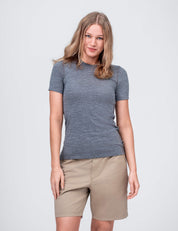 A woman with light brown hair wears the T-shirt Active – Classic Merino 160 Women in grey melange and beige shorts, standing against a plain light background. She smiles slightly, enjoying the T-shirt’s comfortable temperature regulation.