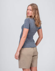 A woman with shoulder-length blonde hair models the T-shirt Active – Classic Merino 160 Women and beige shorts, standing sideways and looking over her shoulder against a plain light background, highlighting temperature regulation.
