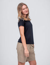 A woman with light brown hair smiles while standing sideways, wearing the T-shirt Active – Classic Merino 160 Women in black and khaki shorts, set against a plain light gray background.