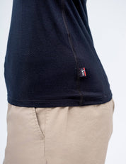 A person wears the T-shirt Active – Classic Merino 160 Women, a lightweight merino tee with a small red and white side seam tag, paired with light beige pants. The plain background highlights the T-shirt’s temperature regulation benefits.