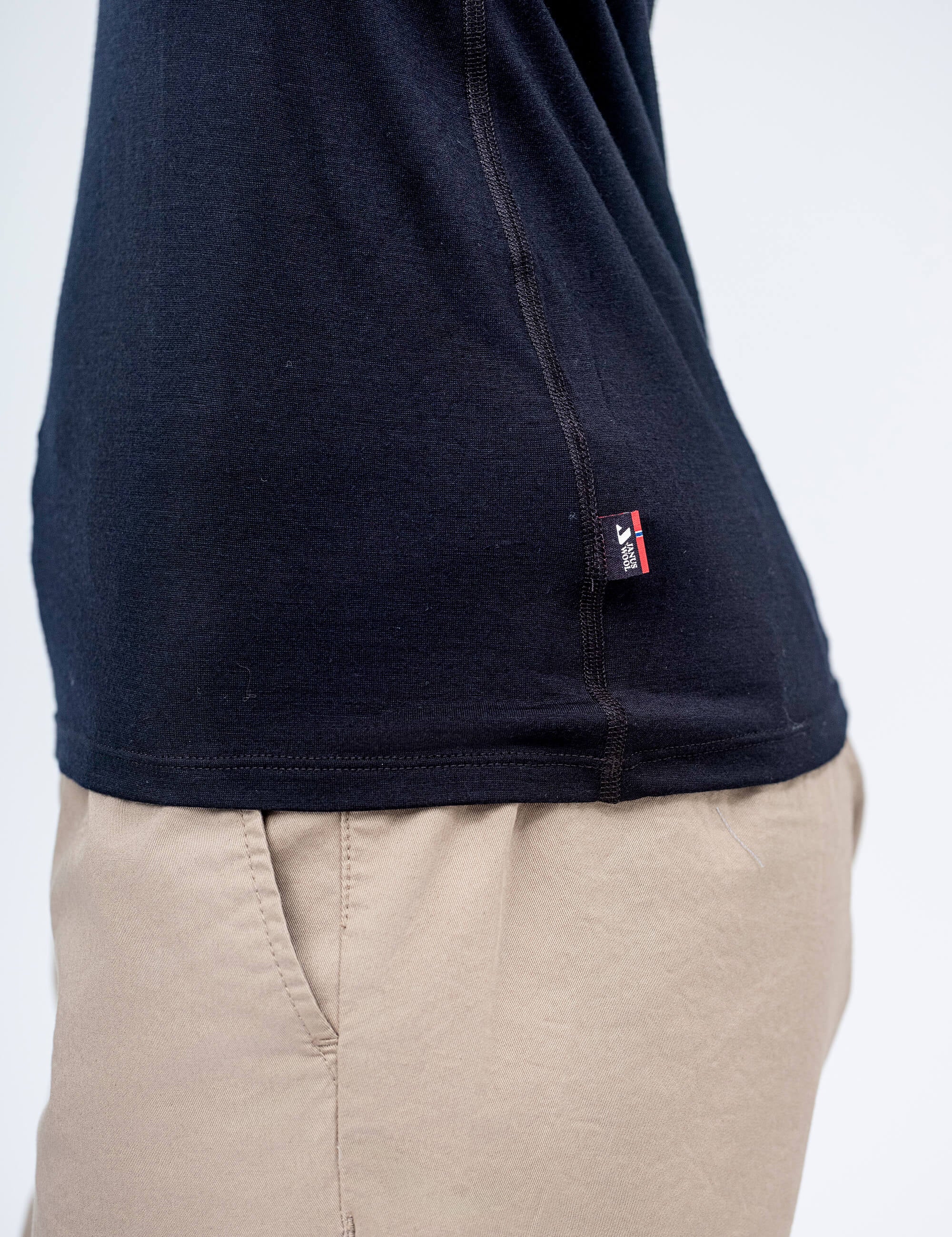 A person wears the T-shirt Active – Classic Merino 160 Women, a lightweight merino tee with a small red and white side seam tag, paired with light beige pants. The plain background highlights the T-shirt’s temperature regulation benefits.