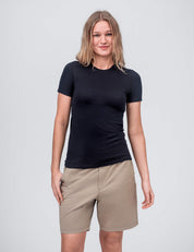 A woman with shoulder-length blonde hair wears the T-shirt Active – Classic Merino 160 Women in black, standing against a plain light gray background and smiling slightly at the camera.