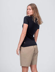 A woman with long, light brown hair models the T-shirt Active – Classic Merino 160 Women and tan knee-length shorts against a plain light gray background, looking over her shoulder.