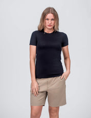 A woman with straight, shoulder-length hair wears the fitted black T-shirt Active – Classic Merino 160 Women and beige shorts, standing against a plain light gray background with one hand in her pocket. The merino T-shirt regulates temperature well.
