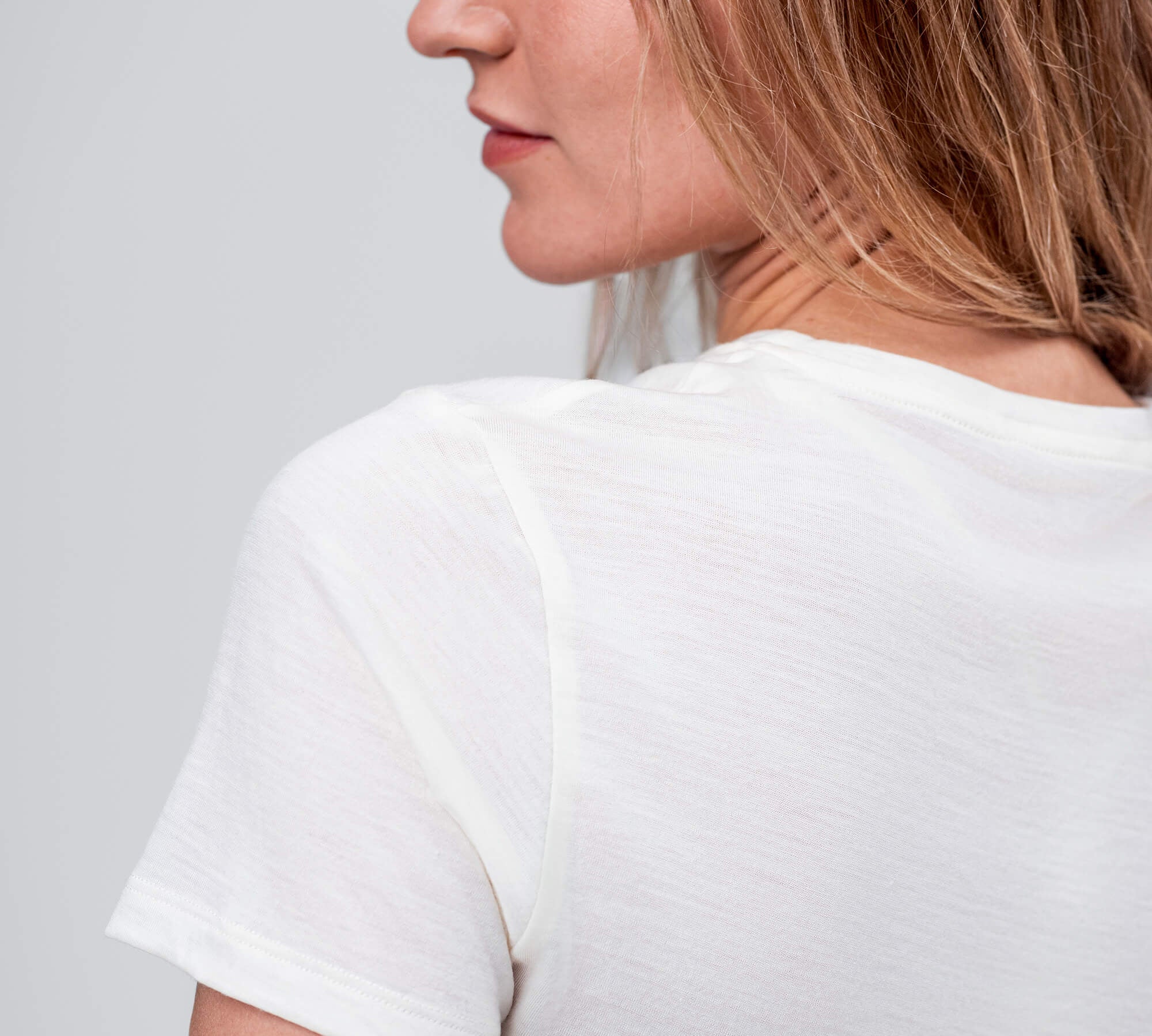 A woman with light brown hair wears the T-shirt Regular – Classic Merino 160 Women in white, seen from the back and side against a plain gray background. The merino fabric ensures breathable, temperature-regulating comfort.