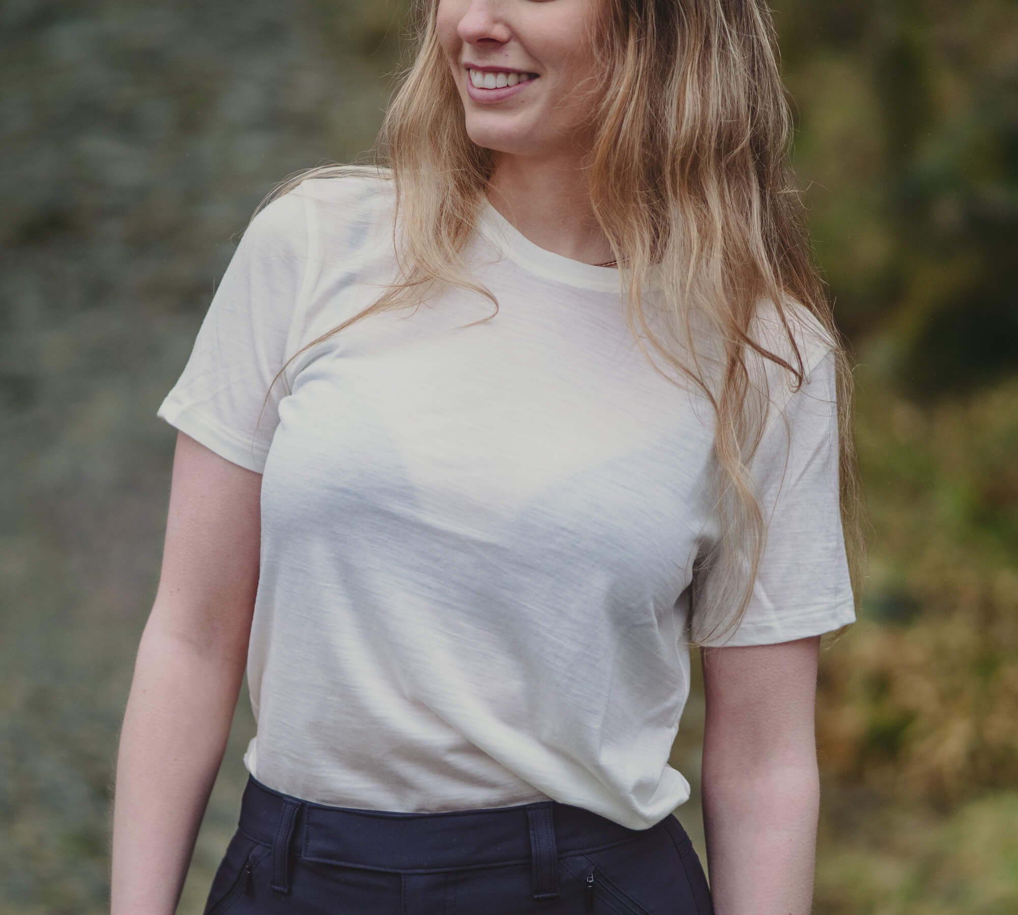 A woman with long, wavy blond hair smiles outdoors among greenery, wearing dark pants and the T-shirt Regular – Classic Merino 160 Women in white.