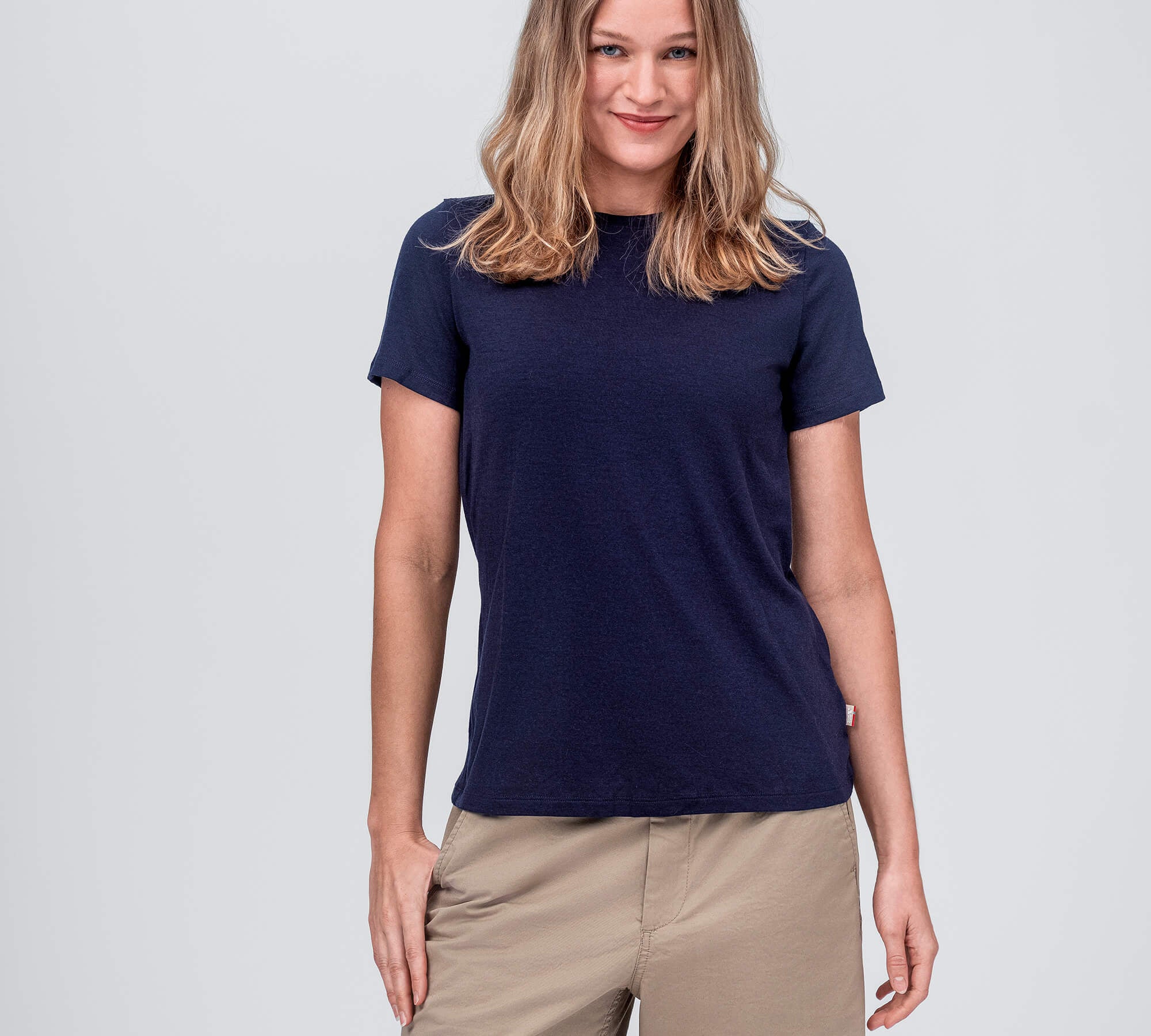 A woman with wavy, shoulder-length hair wears the T-shirt Regular – Classic Merino 160 Women in navy, paired with beige shorts. She stands against a plain light gray background and smiles slightly.