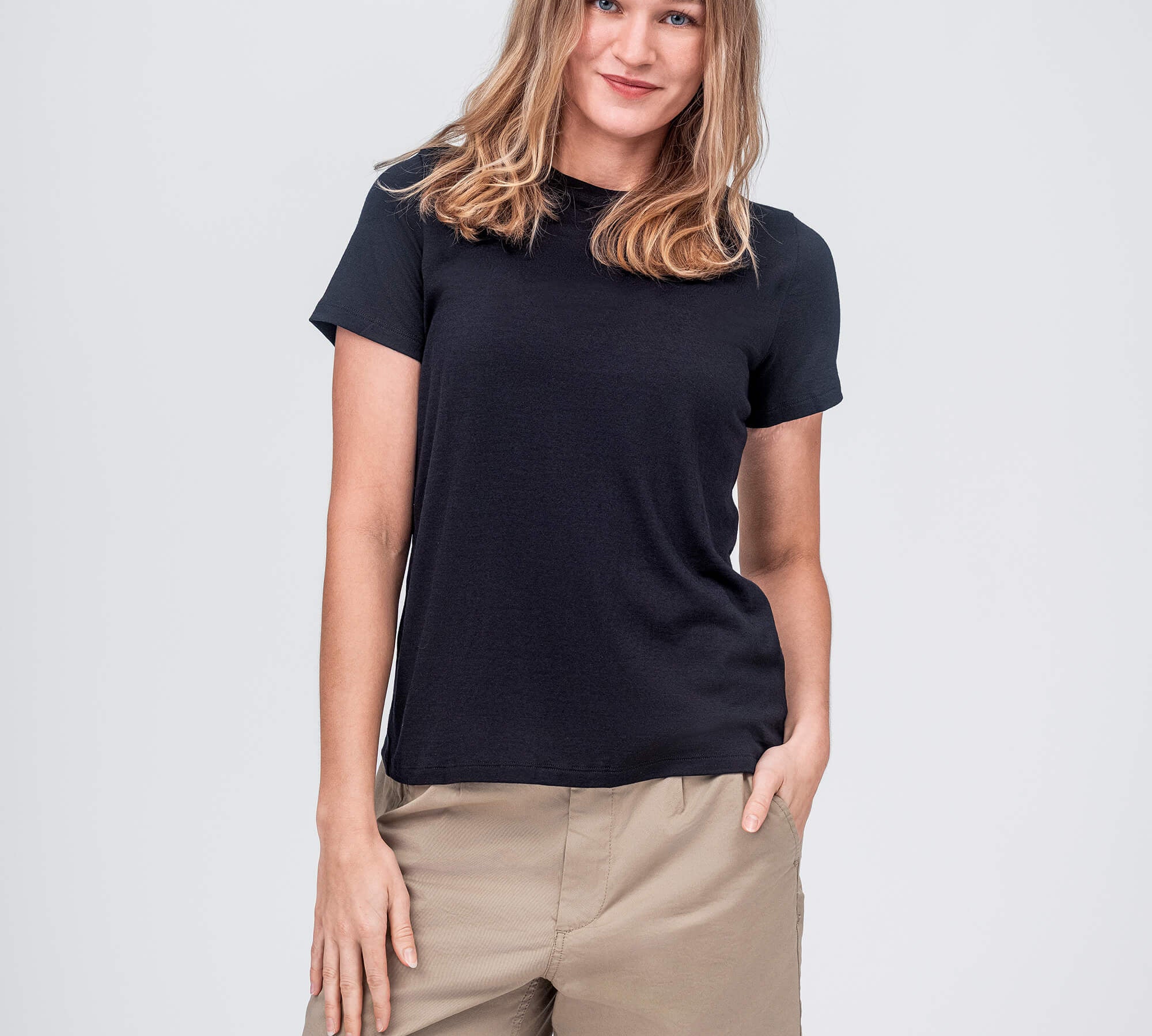 A woman with light brown hair wears the T-shirt Regular – Classic Merino 160 Women in black and beige shorts, standing against a plain light gray background, smiling slightly with one hand in her pocket.