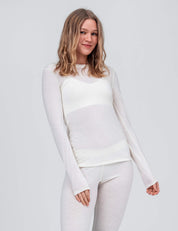 A woman with long, wavy blonde hair models the Classic Merino 160 Women Crew Neck and matching bottoms in Marshmallow, standing against a light grey background and smiling slightly at the camera. Ideal for everyday wear.