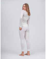 A woman stands barefoot on a light grey background, smiling over her shoulder and wearing the Crew Neck – Classic Merino 160 Women as a fitted, long-sleeve top with matching white leggings—perfect for everyday wear.